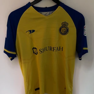 Al Nassr Ronaldo 7 fotbollströja gul/blå - Snygg Al Nassr fotbollströja med Ronaldos namn och nummer 7 på ryggen. Tröjan är gul med blå ärmar och detaljer, har klubbmärke och sponsortryck framtill. Tillverkad i lätt och ventilerande material, perfekt för match eller träning.
