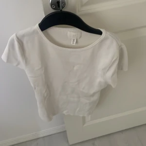 Vit kortärmad topp från H&M - Enkel och stilren vit topp från H&M med rund halsringning och korta ärmar. Perfekt att matcha med jeans eller kjol för en clean look. Gjord i mjuk bomull som känns skön mot huden.