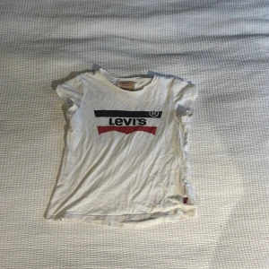 Vit Levi's t-shirt med logga - Vit t-shirt från Levi's med klassisk logga i svart och rött framtill. T-shirten har rund halsringning och korta ärmar. Tillverkad i mjuk bomull och har en avslappnad passform. Perfekt för en chill och stilren look.