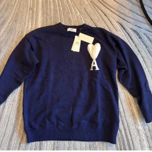 Ami Paris stickad tröja - Snygg mörkblå stickad tröja från Ami Paris med vit hjärtlogga och bokstaven A på bröstet. Klassisk rund halsringning och långa ärmar. Perfekt för dig som gillar stilrena plagg med en twist. Ribbstickade muddar vid ärmslut och nederkant.