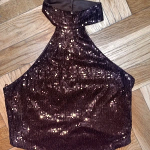 Brun paljett-tubtopp från Gina Tricot - Säljer en brun glittrig tubtopp från Gina Tricot med paljetter över hela framsidan. Toppen har hög hals och knytband i ryggen för en snygg passform. Perfekt för dig som vill sticka ut med en festlig och trendig look.