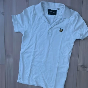 Vit pikétröja från Lyle & Scott: Storlek xs mer som S! - Vit pikétröja från Lyle & Scott med klassisk krage och korta ärmar. Tröjan har det ikoniska broderade guldfågel-loggan på bröstet och är tillverkad i bomull. Perfekt för en clean och stilren look.