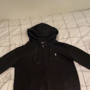 Svart hoodie från Polo Ralph Lauren - Svart hoodie från Polo Ralph Lauren med dragkedja och huva. Klassisk design med vit broderad logga på bröstet och snörning i huvan. Perfekt för dig som gillar en stilren och enkel look. Långärmad och i mjukt bomullsmaterial.