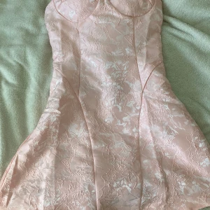 Ljusrosa kortklänning från Peppermayo - Superfin ljusrosa kortklänning från Peppermayo med blommigt mönster och snygg snörning i ryggen. Klänningen har en figurnära passform och är gjord i ett glansigt material som ger en lyxig känsla. Perfekt för dig som vill sticka ut lite extra!