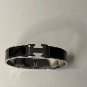 Svart H-armband i metall - Stilrent armband från  med ikoniskt H-spänne i silverfärgad metall och svart emalj. Bred, rund form som sitter snyggt på handleden och ger en lyxig känsla. Perfekt accessoar för dig som gillar klassiska designer och statement-smycken.