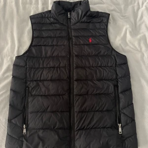 Svart dunväst från Polo Ralph Lauren - Snygg svart dunväst från Polo Ralph Lauren med horisontella quiltade sömmar och hög krage. Västen har två dragkedjefickor framtill och stängs med en dragkedja. Klassisk röd Polo-logga broderad på bröstet. Perfekt för lager-på-lager-stil. En fantastisk fin väst som är använd max 5 gånger. Inga fläckar eller hål eller någonting, den är precis som att den är helt ny. Skulle säga att den passar xs i herr och barn xl, runt 160-180 cm. Hör av er vid frågor! 
