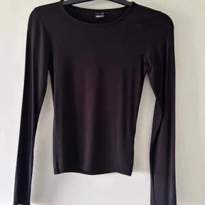 Svart långärmad topp Gina Tricot XS - En svart, enkel och stilren långärmad topp från Gina Tricot i storlek XS. Modellen är figurnära med rund halsringning och mjukt, stretchigt material som sitter snyggt på kroppen. Perfekt att styla med jeans eller kjol för en clean look.