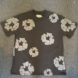 Svart t-shirt med gråa blommor Denim Tears - Svart t-shirt från Denim Tears med unikt grått blommigt mönster över hela plagget. Klassisk passform med rund hals och korta ärmar. Tillverkad i 100% bomull för en skön känsla och snygg streetstil. Det står att det är storlek S men den är väldigt stor så skulle säga att den är storlek M