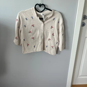 Beige kofta med broderade blommor från Arket - Superfin och söt beige kofta/kortärmad stickad tröja med broderade små blommor i rosa och blått. Koftan har trekvartsärm, knappar framtill och är stickad i mjukt material. Koftan är lite croppad