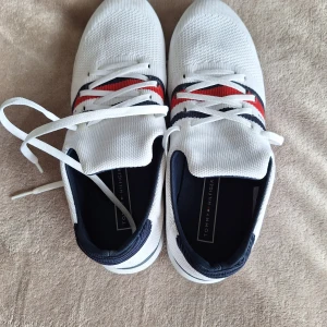 Vita sneakers från Tommy Hilfiger - Snygga vita sneakers från Tommy Hilfiger med stickad ovandel och klassiska röda och marinblå ränder på sidan. Skorna har vita snören, rund tå och detaljer i marinblått vid hälen. Perfekta för en clean och sportig look.