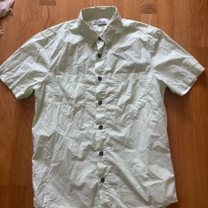 Ljusgrön kortärmad skjorta Stone Island - Säljer en ljusgrön kortärmad skjorta från Stone Island. Skjortan har svarta knappar med logga, två bröstfickor och klassisk krage. Tillverkad i bomull, perfekt för varmare dagar. Enkel och stilren design som passar till mycket.