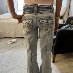Ljusblå bootcut lågmidjade jeans - Säljer ett par ljusblå jeans med bootcut passform. Jeansen har snygga detaljer som dragkedjor och knappar framtill samt broderade bakfickor. Materialet är klassisk denim och modellen har låg midja. Dem har en trasig dragkedja men man kan lätt sy på en ny🩷