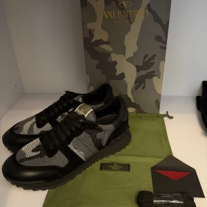 Valentino Garavani Rockrunner sneakers - Snygga Valentino Garavani Rockrunner sneakers i svart och grått med camouflagemönster. Skorna har svarta mockadetaljer, meshpaneler och coola nitar på hälen. Klassisk snörning och rund tå. Perfekta för dig som vill sticka ut med stil. OBS ej använda (helt nya endast testade). Pris kan diskuteras. Storlek 45