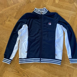 Blåvit track jacket från FILA, M - Snygg blå och vit track jacket från FILA med klassiska ränder på krage och muddar. Jackan har dragkedja framtill, två fickor och broderad FILA-logga på bröstet. Perfekt för en sportig och retro vibe. 
