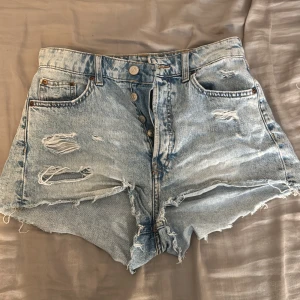 Ljusblå slitna jeansshorts - Säljer ett par ljusblå jeansshorts med hög midja och råa, fransiga kanter. Shortsen har flera slitningar och knappar framtill för en cool, avslappnad look. Perfekta för sommardagar och festivaler.
