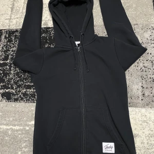 Svart hoodie med dragkedja från Andy by Frank Dandy - Svart hoodie från Andy by Frank Dandy i storlek XS. Tröjan har huva med snörning, hel dragkedja framtill och två fickor. Mjuk och fleeceliknande insida som gör den extra skön. Perfekt för chill dagar eller när du vill ha något bekvämt.