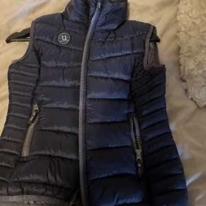 Mörkblå dunväst från Uhip - Snygg mörkblå dunväst från Uhip med hög krage och två dragkedjefickor framtill. Västen har quiltad design och Uhip-logga på bröstet. Perfekt för lager-på-lager och håller dig varm under kyliga dagar. För häst intresserade. Strl 150/xs. Har inte använt mycket endast ca 3 gånger. Har inga defekter