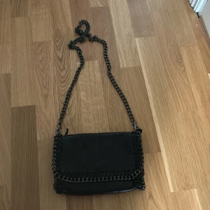 Svart axelväska med kedjedetaljer - Snygg svart axelväska i skinnimitation med grov silverfärgad kedja som axelrem och runt locket. Väskan har lock med magnetknäppning och dekorativ söm längs kanterna. Perfekt storlek för det viktigaste och ger en cool vibe till outfiten.