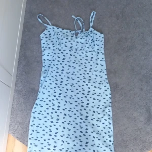 Blommig ljusblå klänning Stories S - Supersöt ljusblå kortklänning från Stories med mörkblått blommönster. Klänningen har smala axelband med knyt och en lätt ribbad struktur i tyget. Perfekt för varma dagar och ger en fräsch sommarkänsla.