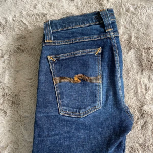 Blå jeans från Nudie Jeans Co - Snygga blå jeans från Nudie Jeans Co med klassisk orange söm och ikonisk broderad bakficka. Jeansen har normal passform och raka ben, tillverkade i slitstarkt bomullsjeans. Perfekta för dig som gillar stilrena och tidlösa jeans.