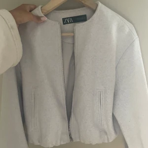 Ljusgrå bomberjacka från Zara XS - Snygg ljusgrå bomberjacka från Zara i storlek XS. Jackan har minimalistisk design, rund halsringning och dragkedja framtill. Tillverkad i mjukt material med två fickor på sidorna. Perfekt för dig som gillar stilrena och enkla plagg.
