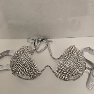 Silvrig bikini med nitar och strass - Supersnygg bikiniöverdel i metallic-silver med massor av glittrande strass och coola silverfärgade nitar. Knytband i nacken och ryggen för perfekt passform. Perfekt för dig som vill sticka ut på stranden eller poolpartyt.