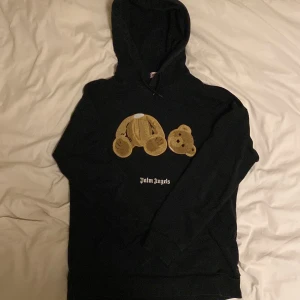 Svart Palm Angels hoodie med björn - Svart hoodie från Palm Angels med ikoniskt björnmotiv på bröstet och logga under. Tröjan har huva, långa ärmar och är tillverkad i mjuk bomull. Perfekt för dig som gillar streetwear och vill sticka ut med en unik detalj framtill.