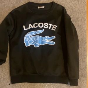 Svart sweatshirt från Lacoste med blå logga - Använt ett par fåtal gånger. Svart sweatshirt från Lacoste med stor blå krokodil och vit Lacoste-text på bröstet. Tröjan har rund halsringning och långa ärmar. Perfekt för dig som gillar streetstyle och vill sticka ut med en ikonisk logga.
