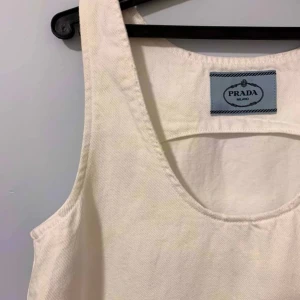 Vit bomullsklänning från Prada - Superfin vit klänning från Prada i 100% bomull. Ärmlös modell med rund halsringning och lätt A-linjeformad siluett. Enkel och stilren design, perfekt för dig som gillar minimalistiskt mode. Tillverkad i Italien och har diskret Prada-logga vid halsen.