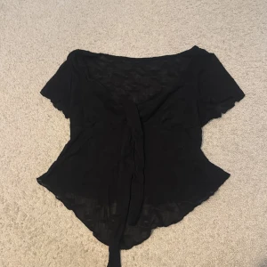 Svart omlottblus med knyt - Säljer en svart omlottblus med korta ärmar och knyt framtill. Toppen har en lätt och luftig känsla med v-ringad skärning och asymmetrisk nederkant. Perfekt för dig som gillar en stilren och enkel look.