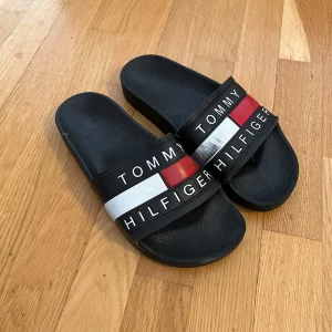 Svarta sandaler  - Svarta sandaler  med bred rem över foten i svart, vitt och rött. Logotypen är tryckt på remmen. Perfekta för sommaren och chill dagar. Tillverkade i syntetmaterial med platt sula och öppen tå.