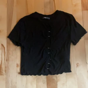Svart croppad topp med knappar - En svart croppad topp med korta ärmar och dekorativa knappar framtill. Toppen är helt öppen vid ringarna mitt på tröjan, den har även vågig kant nertill och vid ärmslut. Otroligt skönt och stretchigt material.