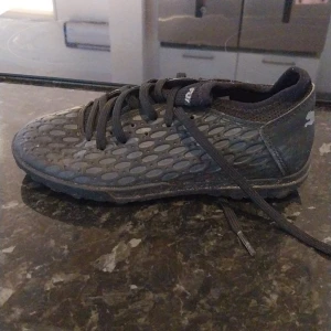 Svarta fotbollsskor från Puma - Säljer ett par svarta fotbollsskor från Puma med coolt mönstrad ovansida och klassisk logga på hälen. Skorna har snörning och en låg profil, perfekt för dig som vill ha stilrena och sportiga skor på planen.