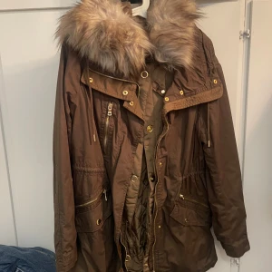 Brun parkajacka med fuskpäls från Mango - Snygg brun parkajacka från Mango med stor fluffig fuskpäls på huvan, guldfärgade dragkedjor och knappar. Jackan har flera fickor, justerbar midja och är perfekt för kyliga dagar. Materialet är slitstarkt och jackan har en klassisk parkamodell.