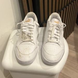 Vita Puma sneakers med gulddetaljer - Fräscha vita Puma sneakers med klassisk låg profil och guldfärgad logga på sidan och tungan. Skorna har snörning, rund tå och är tillverkade i syntetmaterial med detaljer i skinn. Perfekta för en clean och sportig look.