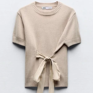 Beige stickad topp med knyt från Zara - Superfin beige stickad topp från Zara med korta ärmar och rund hals. Snygg detalj med knytband i midjan som ger en feminin siluett. Perfekt att styla med jeans eller kjol för en trendig look.