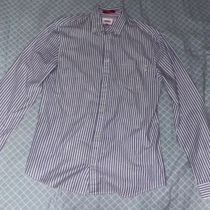 Randig skjorta från Tommy Hilfinger, slim fit - Snygg randig skjorta från Tommy Jeans i slim fit-modell. Skjortan är blå och vit med klassisk krage, bröstficka och knappar framtill. Skriv för fler frågor eller bilder i chatten tack!