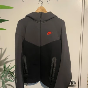 Svart och grå Nike hoodiejacka S - Nike hoodiejacka i svart och mörkgrått med röd logga på bröstet. Jackan har hel dragkedja framtill, två fickor med dragkedja och en skön huva. Tillverkad i mjuk bomullsblandning, perfekt för chill eller träning. båda byxorna och tröjan säljs med varandra.