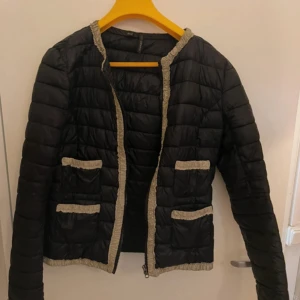 Svart quiltad pufferjacka med beige detaljer - Snygg svart quiltad pufferjacka med beige rynkade detaljer längs kanter och fickor. Jackan har dragkedja framtill, två framfickor och en kortare, boxig passform. Perfekt för dig som vill ha en stilren men unik jacka till vardags.