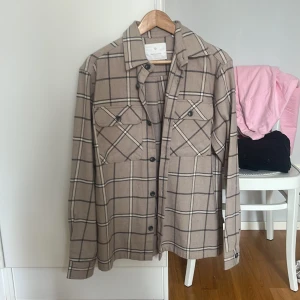 Beige rutig overshirt med knappar - Snygg beige overshirt från Weekday med rutigt mönster i svart och vitt. Skjortan har två bröstfickor med knappar, klassisk krage och långa ärmar. Perfekt att bära öppen eller stängd för en avslappnad stil.