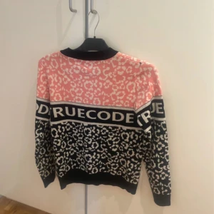 mönstrad tröja från BFashion strl S/M  - Cool stickad tröja från BFashion med rosa och svart mönster och stor text 'TRUECODE' över bröstet. Tröjan har rund halsringning, långa ärmar och är gjord i en mix av ull och syntet. Perfekt för dig som vill sticka ut med en trendig look.