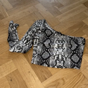 Oneshoulder topp med ormmönster från SHEIN - Säljer en oneshoulder topp från SHEIN med svart, grått och vitt ormmönster. Toppen har lång ärm på ena sidan och ärmlös på andra, vilket ger en cool asymmetrisk look. Perfekt för dig som vill sticka ut med ett djurmönster och edgy stil.