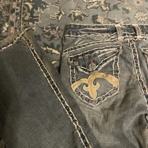 Blå jeans med broderade fickor - Säljer ett par blå jeans från Miss Me med kontrastsömmar och snygga broderade detaljer på bakfickorna. Jeansen har bootcut-modell och coola slitningar för en edgy look. Perfekta för dig som gillar statement-jeans med unika detaljer.