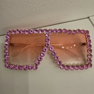 Rosa glittriga solglasögon med stenar - Statement solglasögon med stora fyrkantiga bågar, dekorerade med glittrande rosa stenar runt hela ramen. Glasen är ljusrosa och bågarna är transparenta. Perfekta för dig som vill sticka ut och addera extra bling till din look.