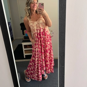 Blommig långklänning i rosa och beige - Superfin långklänning med smala axelband och flowy passform. Klänningen har en beige överdel som övergår i en starkt rosa kjol, båda med blommigt mönster i ljusa toner. Perfekt för varma dagar och ger en riktigt chill vibe.Helt ny.Storlek är märkt som M (plus size) så passar från L till XL