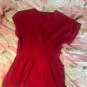 Röd omlottopp med markerad midja - Snygg röd omlottopp från H&M med v-ringning och bred, markerad midja. Toppen har korta ärmar och lätt puffig form nedtill. Perfekt för dig som gillar färg och vill ha en feminin siluett.