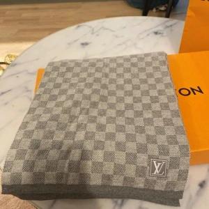 Grå rutig halsduk från Louis Vuitton - Snygg grå halsduk från Louis Vuitton med klassiskt rutigt mönster och diskret LV-logga i ena hörnet. Halsduken är tillverkad i 100% ull och känns både lyxig och stilren. Perfekt accessoar för att lyfta din outfit under kyliga dagar.