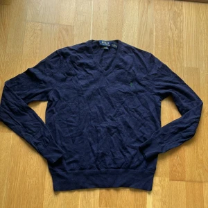 Mörkblå v-ringad tröja Polo Ralph Lauren - Mörkblå v-ringad tröja från Polo Ralph Lauren med klassisk broderad logga på bröstet. Tröjan har lång ärm, ribbade muddar och är tillverkad i mjuk bomull. Perfekt för lager-på-lager och stilren look.