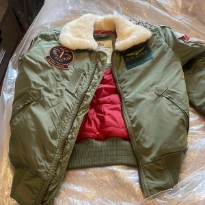 Alpha Industries grön bomberjacka S - Säljer en grön bomberjacka från Alpha Industries med coola patches, röd quiltad insida och vit fuskpäls på kragen. Jackan har dragkedja, ribbade muddar och flera detaljer som ger en riktigt street och militär vibe.