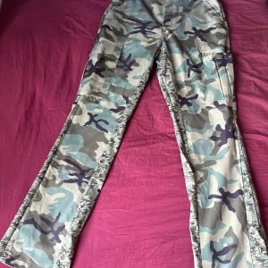 Cargobyxor med camouflage och vida ben - Säljer ett par cargobyxor med grå, blå och gröna camouflage-mönster. Byxorna har vida ben och stora fickor på sidorna. Perfekta för dig som gillar streetstyle och vill sticka ut lite extra. Materialet känns som bomull och de har en loose passform.